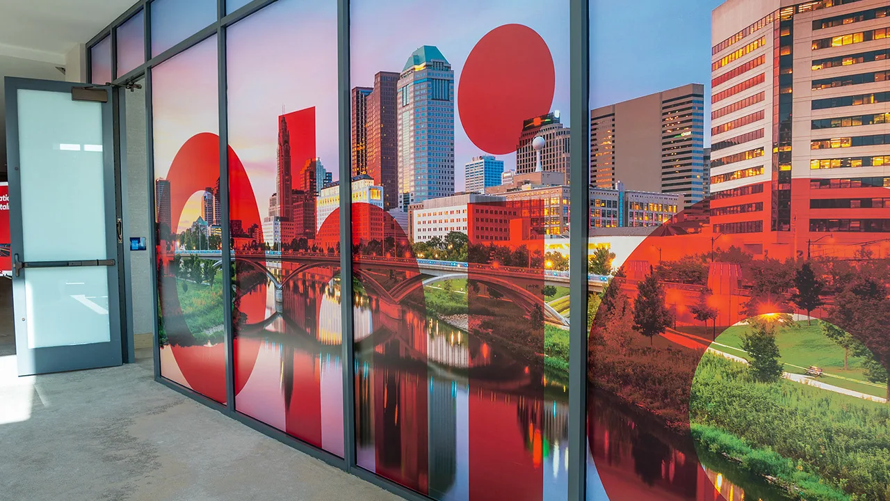 Window film along a glass corridor, the Columbus skyline reflected in the river behind a red sun